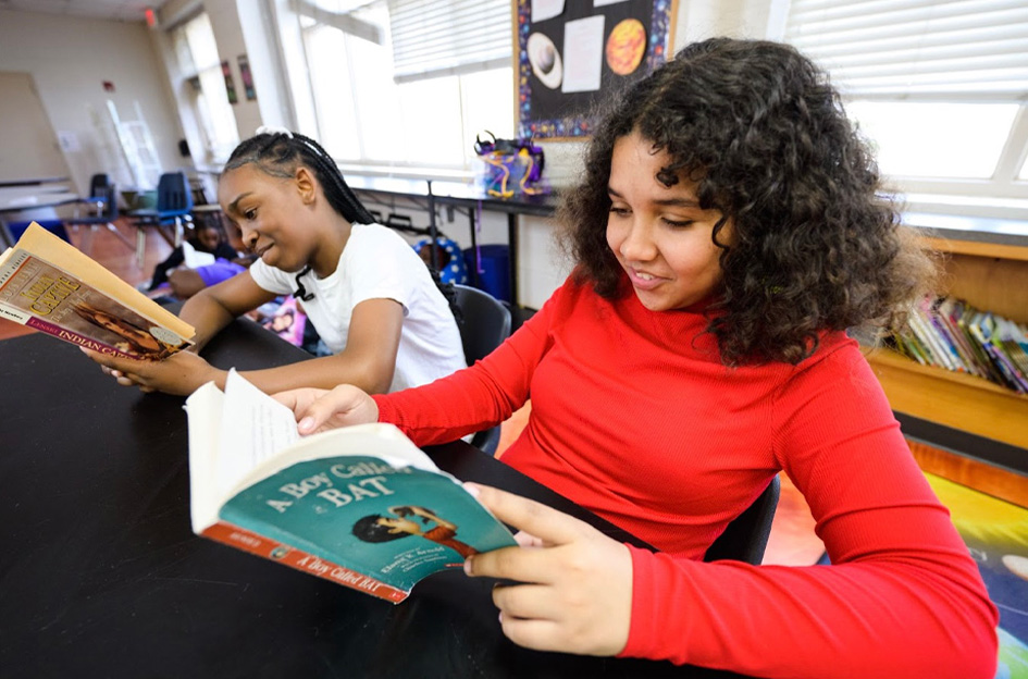 Two smiling girls sitting at a table in a classroom and reading chapter books