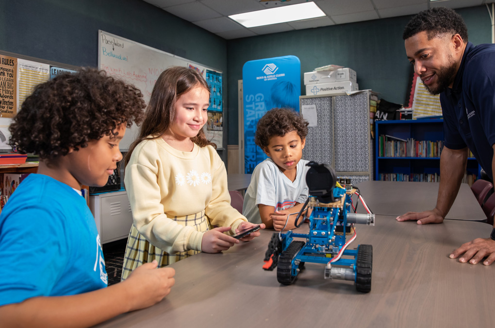 A girl with a smile operates a robot on a table, observed by two boys and a Club mentor, in a classroom.