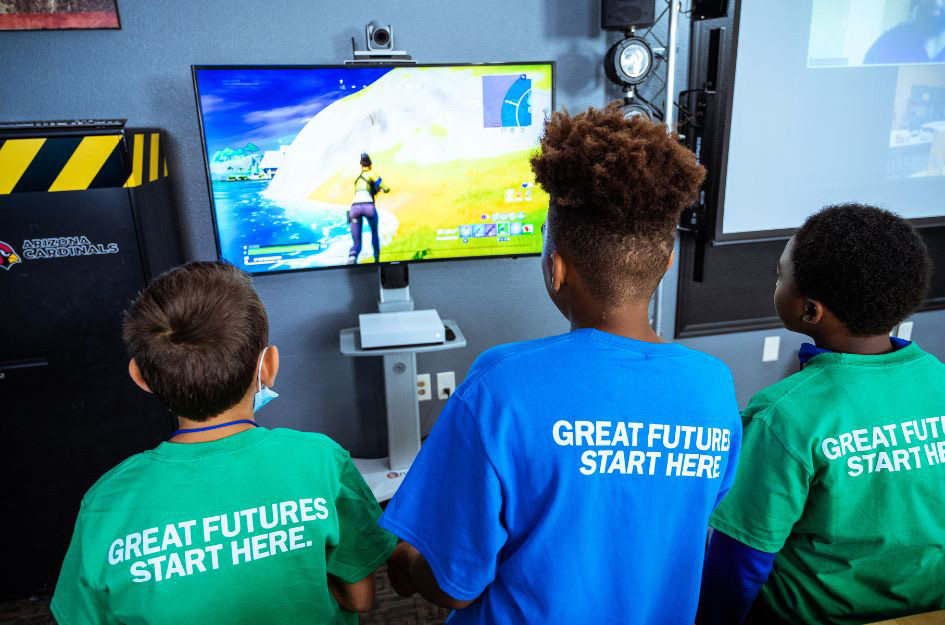 Three Boys &amp; Girls Club members play a video game together, wearing shirts with 'Great Futures Start Here' printed on the back.